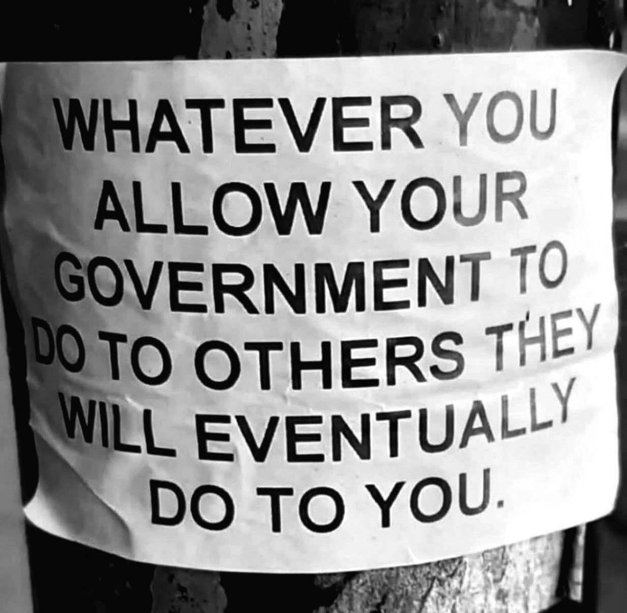 A printed sign reads, WHATEVER YOU ALLOW YOUR GOVERNMENT TO DO TO OTHERS THEY WILL EVENTUALLY DO TO YOU, affixed to a pole.