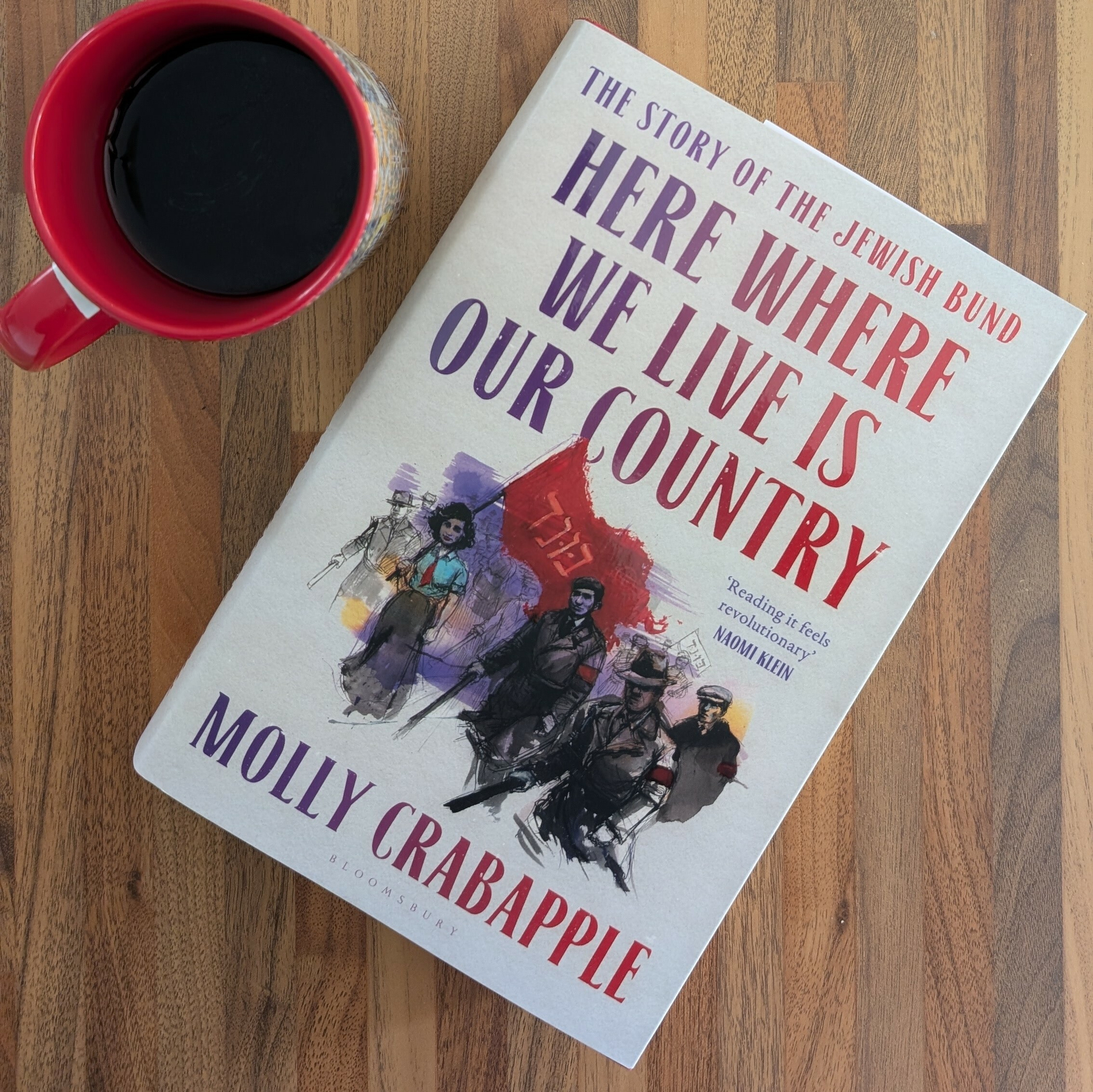 A red mug filled with a dark beverage is next to a book titled Here Where We Live Is Our Country by Molly Crabapple on a wooden surface.