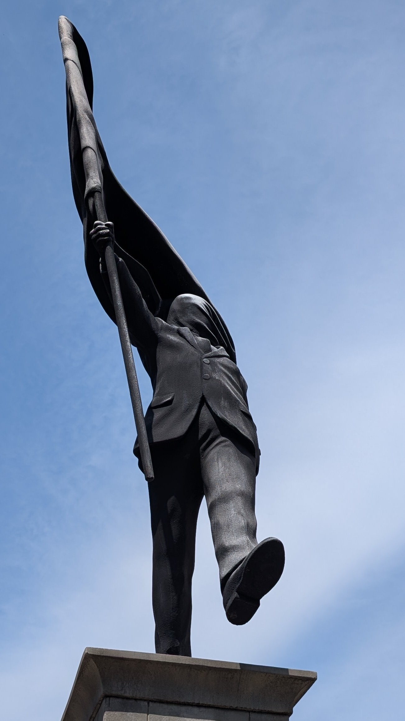 A statue by Banksy depicts a slightly corpulent man in a suit holding a large flowing flag, set against a clear blue sky. The flag covers his face. The pose is as though he is walking ahead. One foot is on the plinth and the other is ahead, nothing underneath as though he is walking off a cliff, blinded by nationalism.